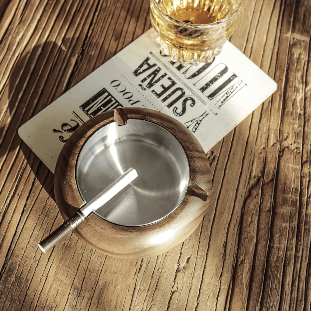 Round wooden ashtray made of walnut with stainless steel liner, placed on a rustic wooden table.