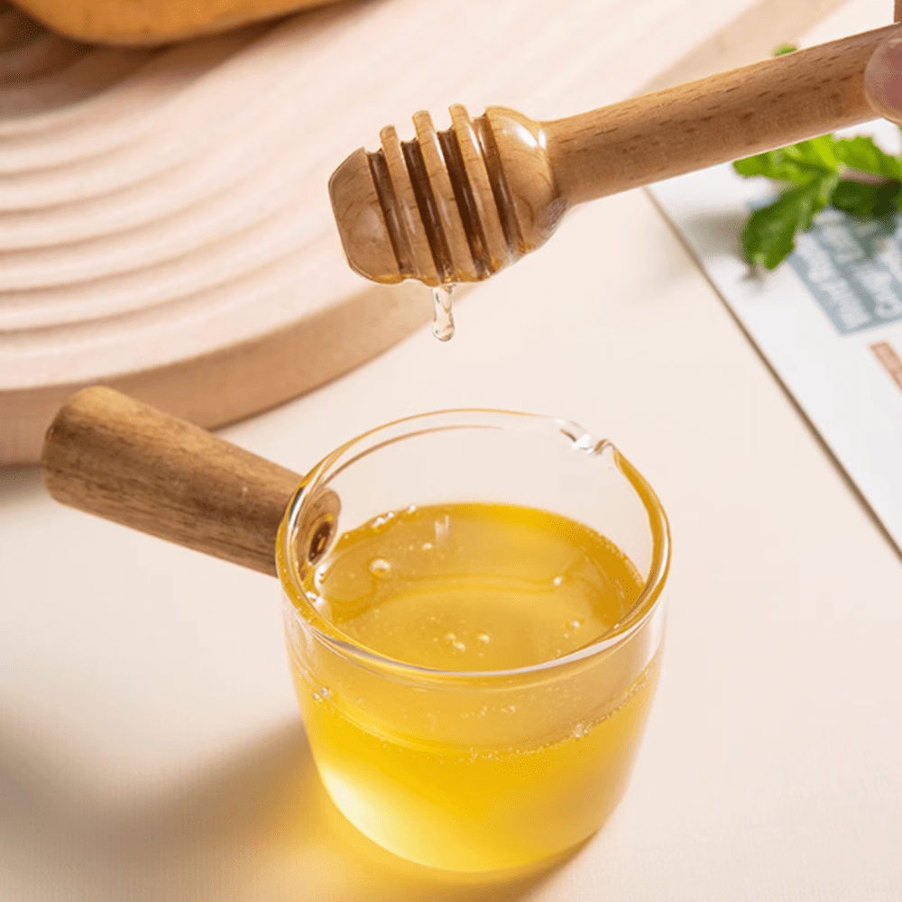 Beechwood stirring stick dripping honey into a glass bowl, showcasing natural texture and design.