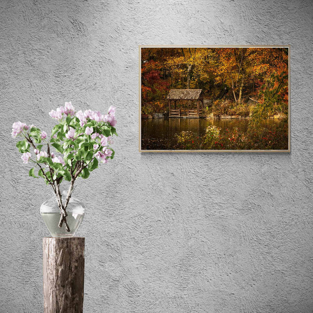 Framed Autumn Lakeside puzzle artwork above a flower vase on a wooden pedestal, showcasing autumn colors.