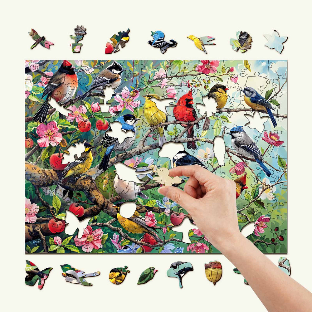 Hand placing a piece in a colorful Birds in the Orchard wooden jigsaw puzzle featuring various birds and flowers.