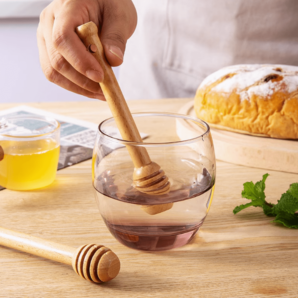 Beechwood stirring stick mixing honey in a glass, accompanied by bread and a jar of honey on a wooden table.