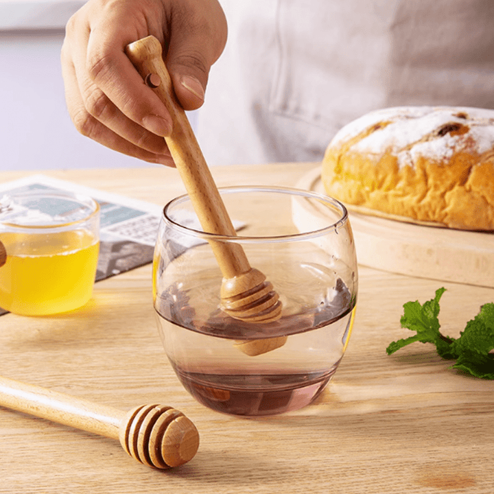 Beechwood stirring stick mixing honey in a glass, accompanied by bread and a jar of honey on a wooden table.