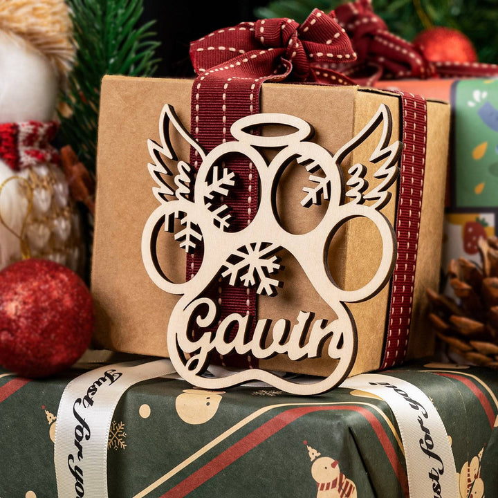 Personalized wooden ornament with paw print and angel wings, featuring name 'Gavin', on wrapped Christmas gift.