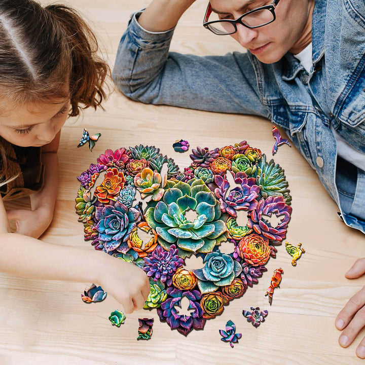 A father and daughter solving a colorful heart-shaped succulent jigsaw puzzle on a wooden table.