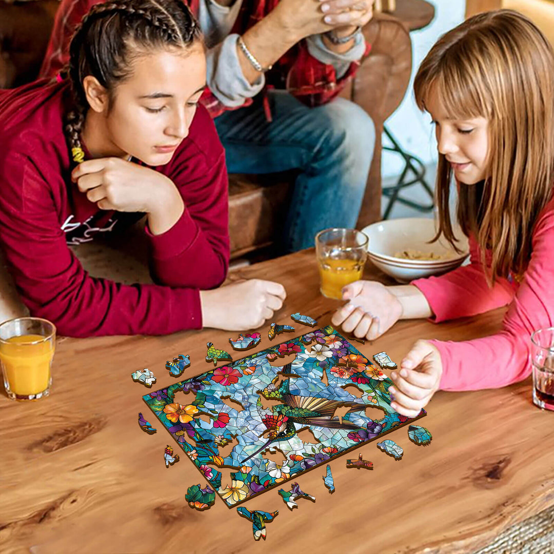 Two girls working on a Stained Glass Hummingbird jigsaw puzzle together at a wooden table.
