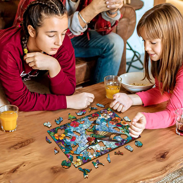 Two girls working on a Stained Glass Hummingbird jigsaw puzzle together at a wooden table.