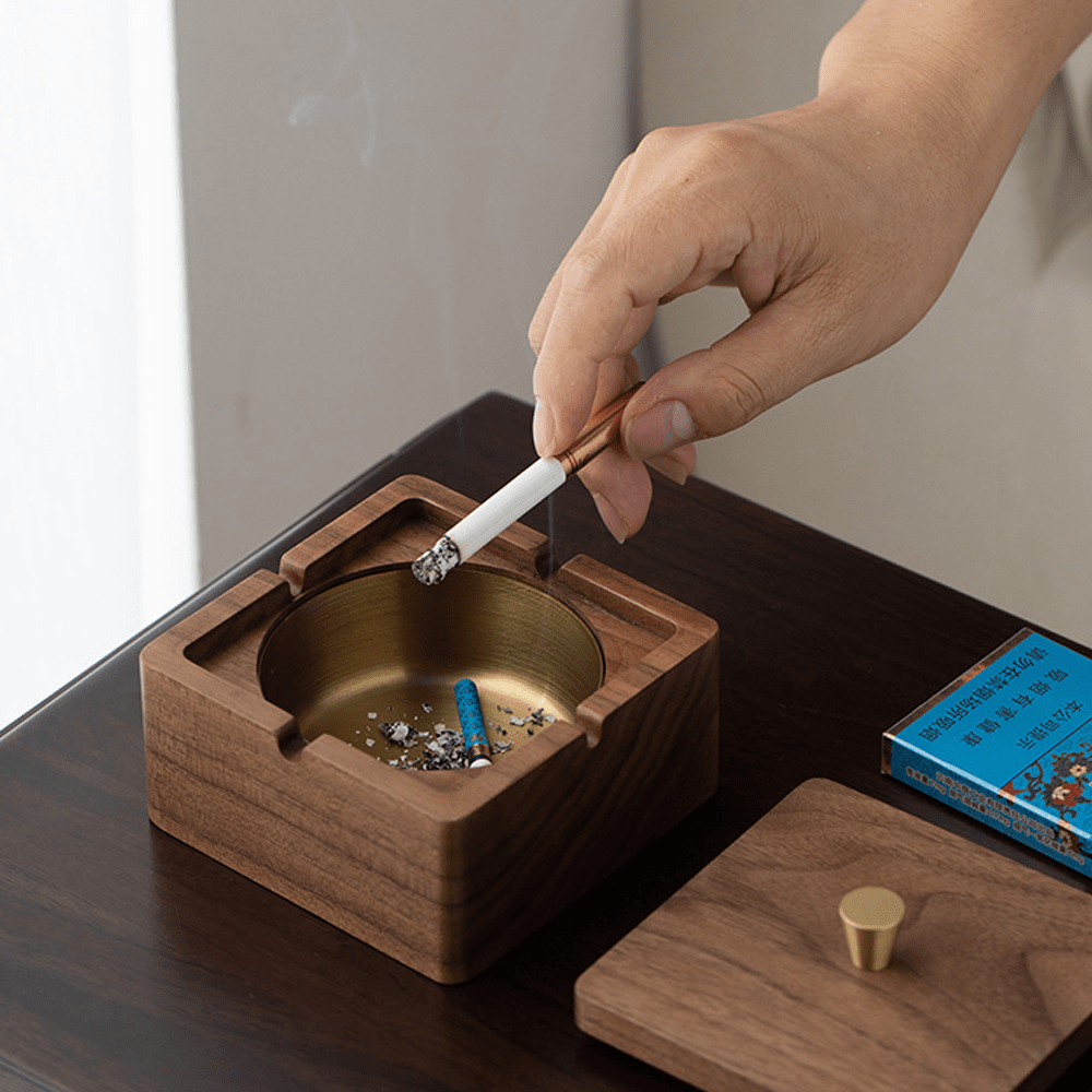 Hand placing a cigarette in a square walnut ashtray with brass liner, showcasing a modern and natural design.