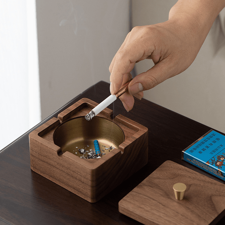 Hand placing a cigarette in a square walnut ashtray with brass liner, showcasing a modern and natural design.