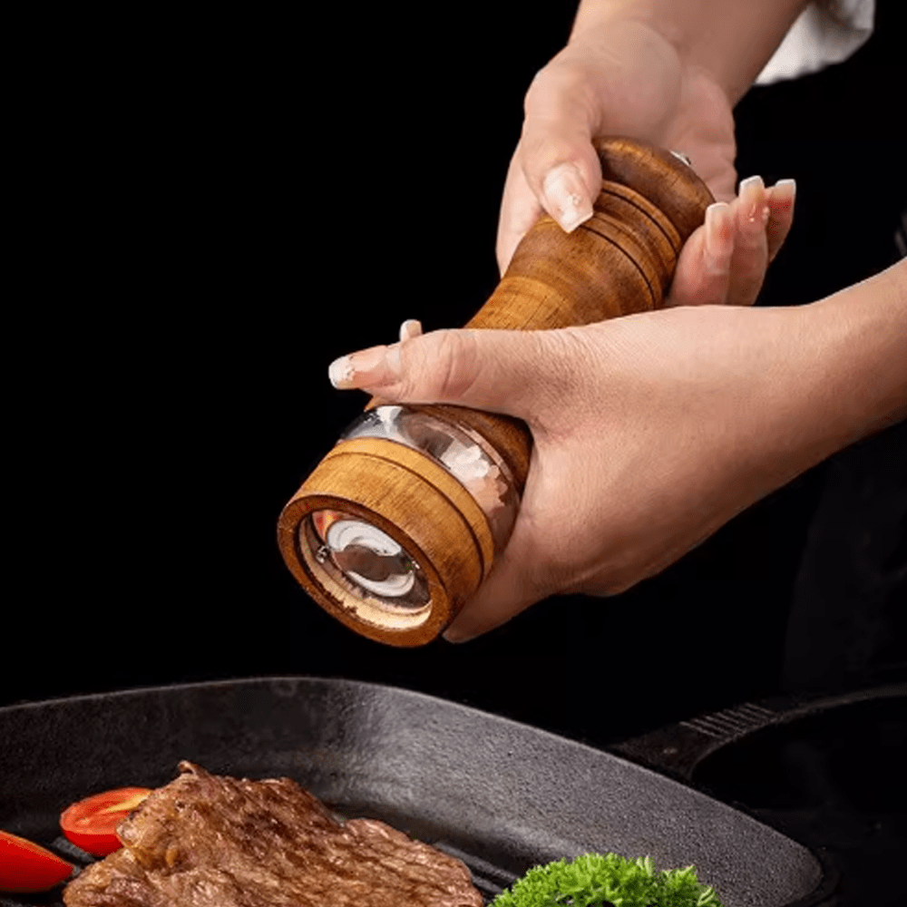 Person using a rubberwood two-tone spice grinder over a sizzling steak, showcasing its unique design and functionality.