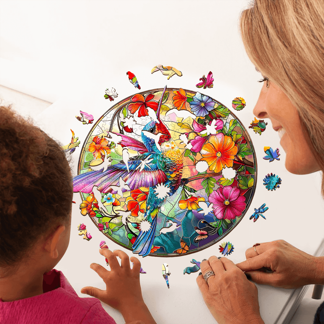 A woman and child engaged in assembling the Stained Glass Hummingbird-2 wooden jigsaw puzzle surrounded by colorful pieces.