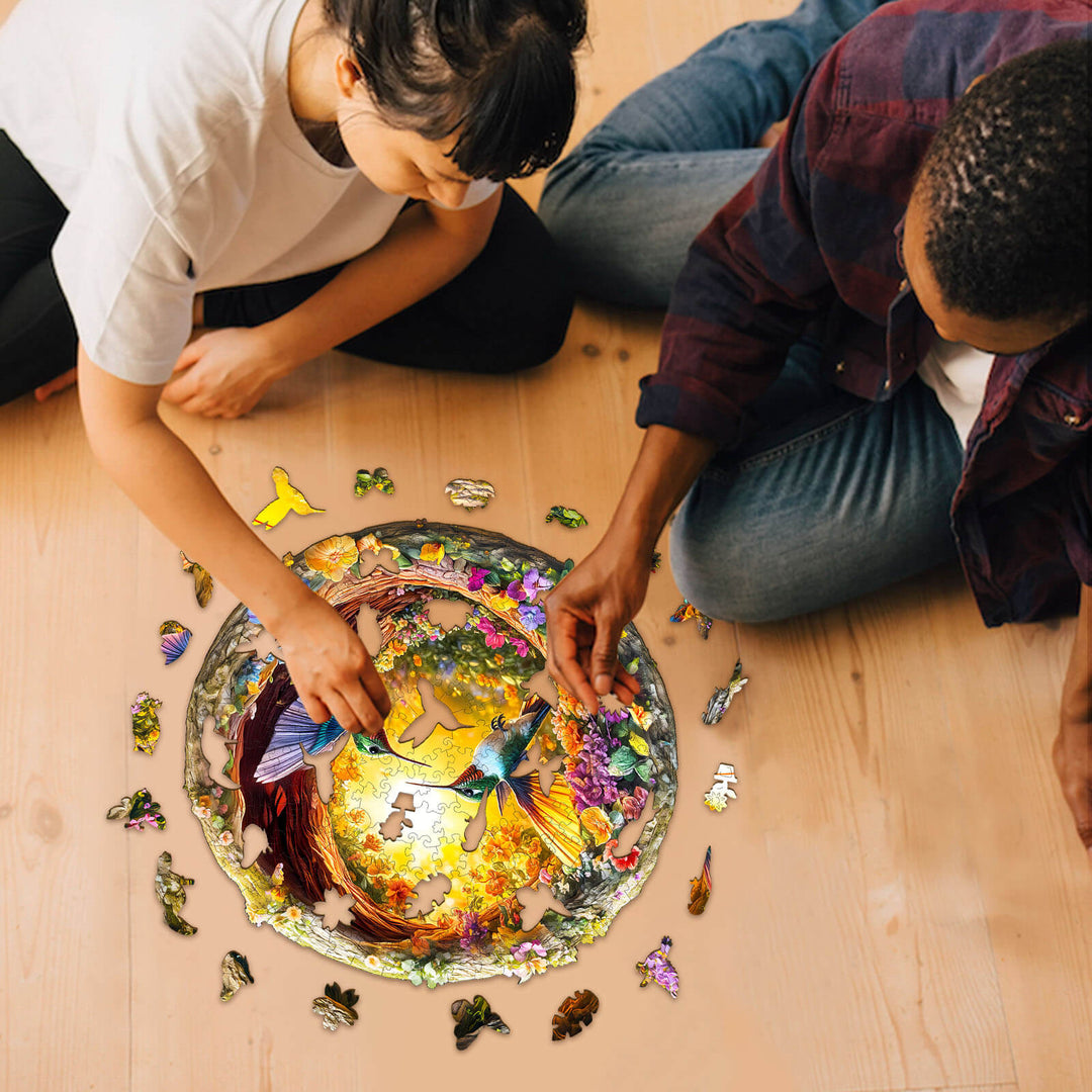 Two people solving the 3D Hummingbird in the Cave-2 wooden jigsaw puzzle on a wooden floor.