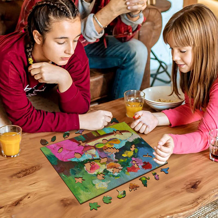 Two girls engaged in a puzzle featuring colorful artwork by Alexander Shandor, surrounded by drinks and puzzle pieces.