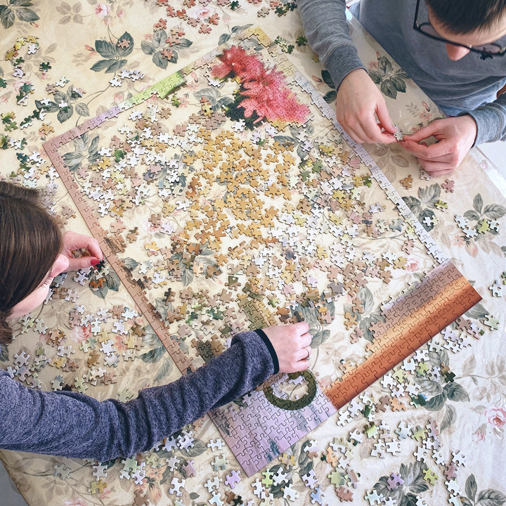 Two people assembling a floral jigsaw puzzle on a table filled with pieces, showcasing a fun, engaging activity.