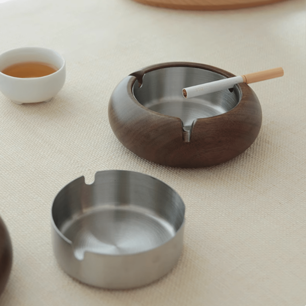 Round wooden ashtray with stainless steel liner and cigarette, alongside a tea cup on a light surface.
