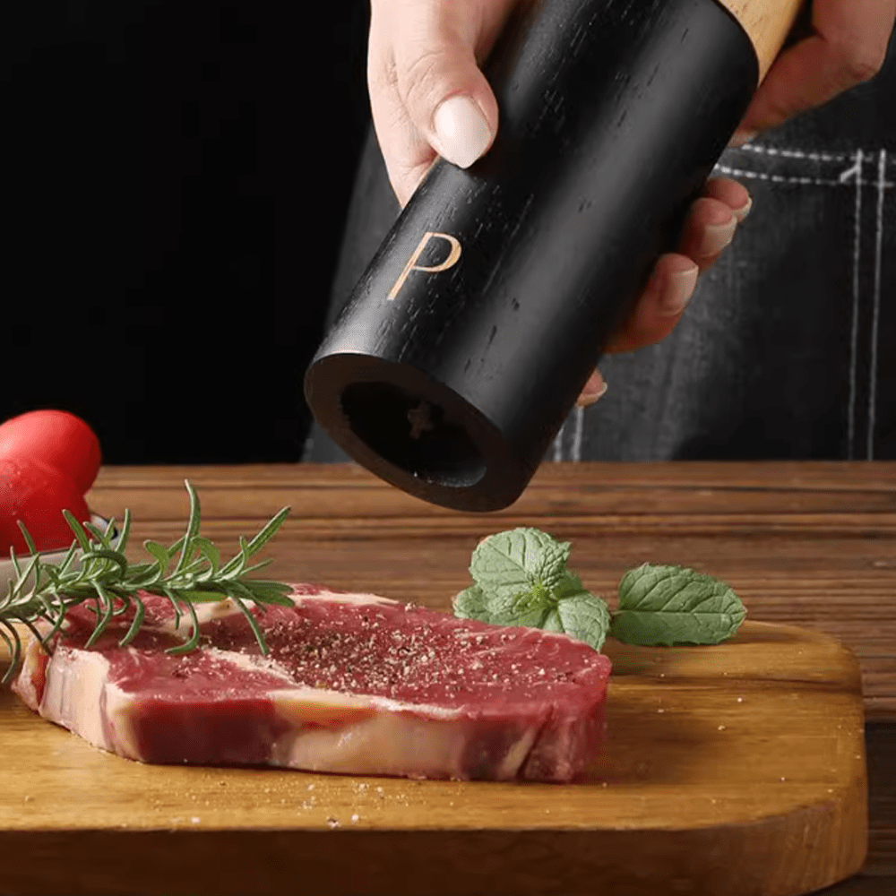 Person using a black pepper grinder to season a steak on a wooden cutting board with herbs.