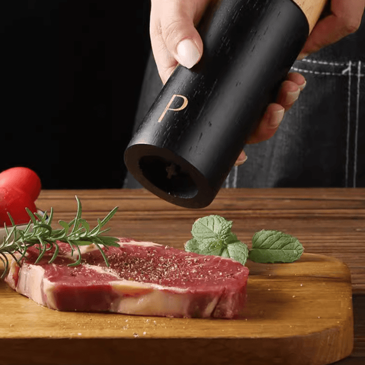Person using a black pepper grinder to season a steak on a wooden cutting board with herbs.