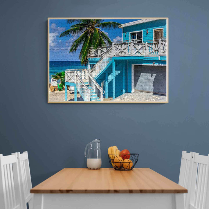 Bright blue beach house with palm trees, framed on a wall above a dining table with fruit and water pitcher.