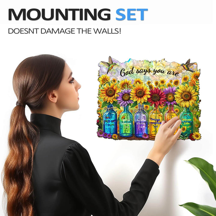 Person using a mounting set to hang a colorful puzzle with sunflowers and the phrase 'God says you are' on the wall.