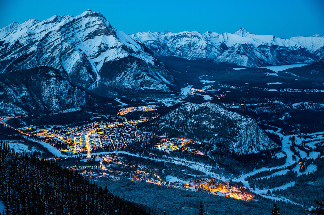 A breathtaking winter landscape at dusk, featuring snowy mountains and a lit valley town nestled among trees.