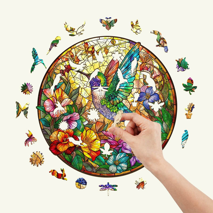 Hand placing a piece in the Stained Glass Hummingbird wooden jigsaw puzzle surrounded by colorful floral designs.