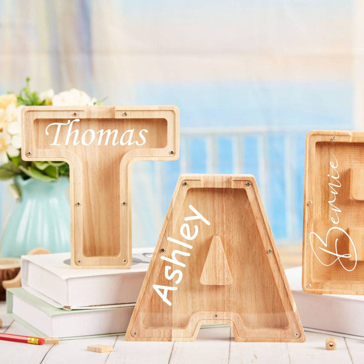 Personalized wooden letter piggy banks with names Thomas, Ashley, and Bernie displayed on a table.