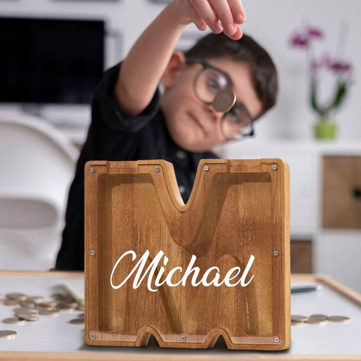 Child dropping a coin into a personalized wooden letter piggy bank with the name Michael.