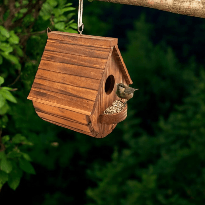 Natural wooden outdoor birdhouse with a bird perched on the feeder, set against green foliage.