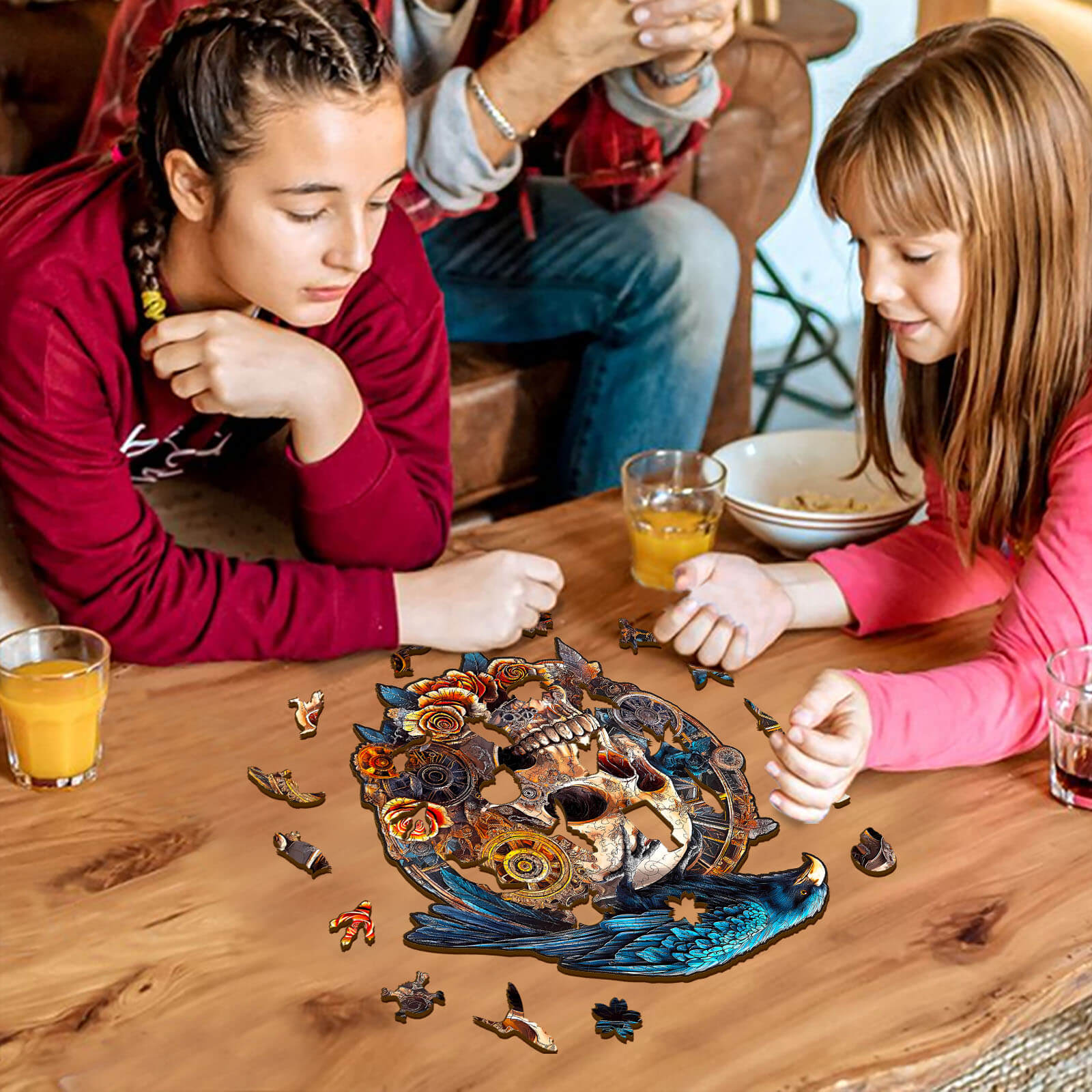 Two girls assembling a 3D Mechanical Skull & Raven wooden jigsaw puzzle on a wooden table with drinks.
