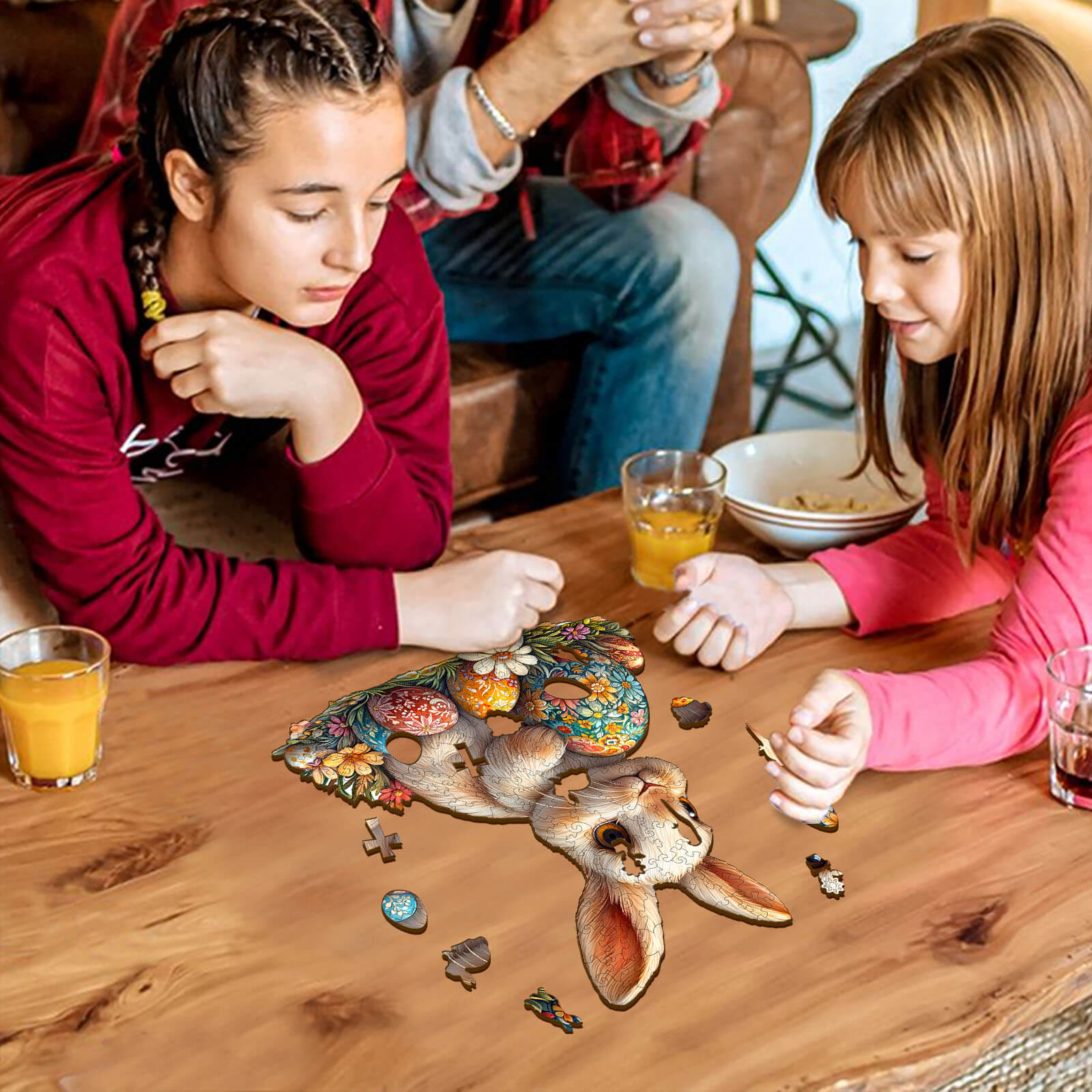 Two children assembling a colorful 3D Easter bunny jigsaw puzzle on a wooden table, surrounded by drinks.