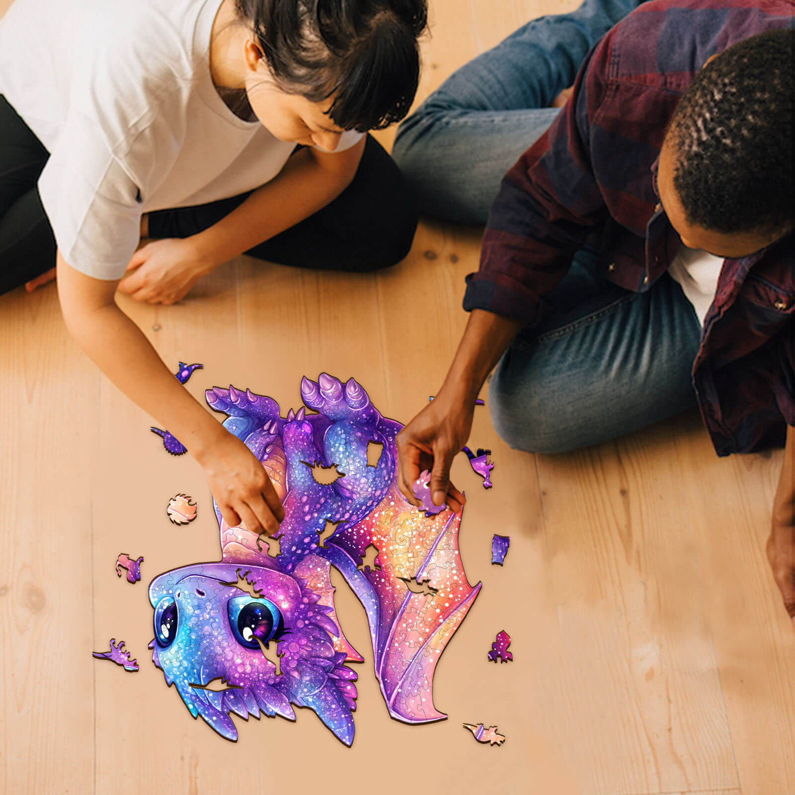 Two people assembling the Glittering Dragon wooden jigsaw puzzle on a wooden floor, showcasing vibrant colors and engaging design.