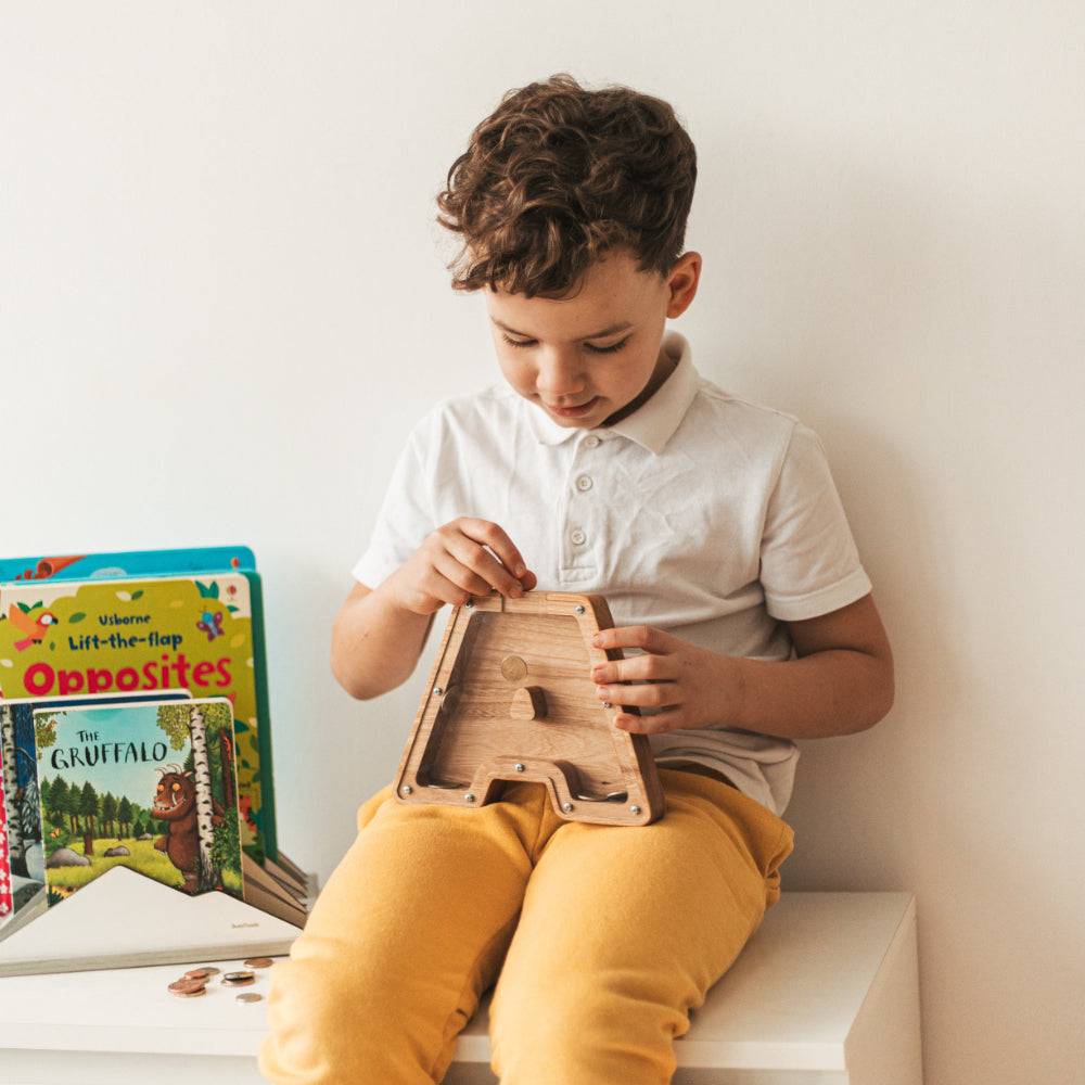 Child using a personalized wooden letter piggy bank, surrounded by colorful books.