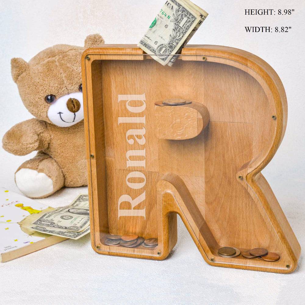 Personalized wooden letter 'R' piggy bank with coins, featuring the name 'Ronald', beside a teddy bear.